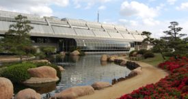 Water runs between a glass-walled complex and a dirt walking path lined with plants and flowers