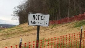 White rectangular sign says "Notice Waters of US" with a tan hill and a row of trees without leaves behind it