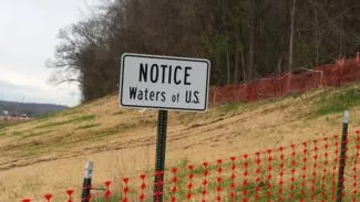 White rectangular sign says "Notice Waters of US" with a tan hill and a row of trees without leaves behind it