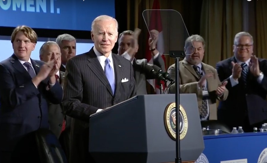 President Joe Biden stands smiling in front of a podium. Five people stand behind him, clapping.