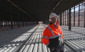 A worker wearing a white hardhat and orange jacket stands smiling atop a metal floor of an under construction factory