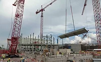 Y12 nuclear clean-up A large red crane lifts a gray rectangular slab with workers wearing yellow vests below, a cloudy sky in the background