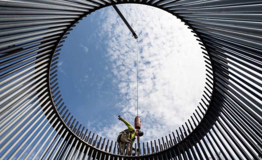 a view from within a circle of bars forming a wind turbine cage, a worker is on top, blue sky with clouds behind