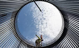 a view from within a circle of bars forming a wind turbine cage, a worker is on top, blue sky with clouds behind