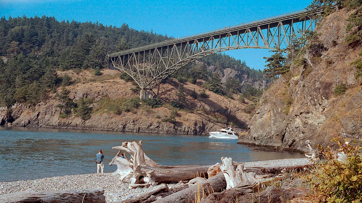 Waters of Puget Sound flow between two mountainous banks with a bridge spanning them and a pine forest beyond.