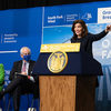 NY Governor Kathy Hochul gestures with her hand as she speaks behind a dais. In the background a large banner notes a 2022 groundbreaking for South Fork Wind Farm, "New York's First Offshore Farm." 