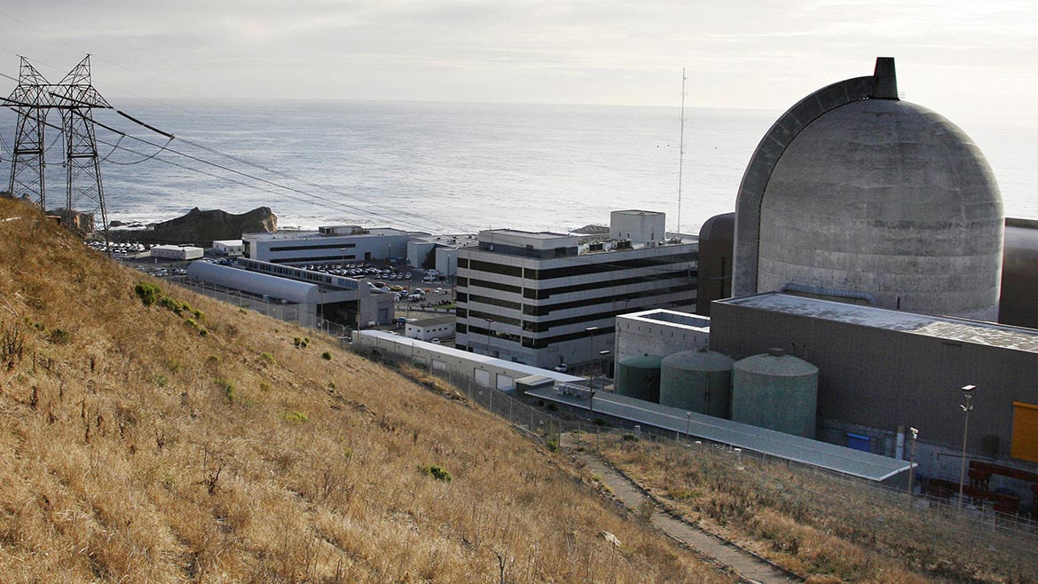 The Diablo Canyon Nuclear Plant, including the domed containment building and transmission line shown. A body of water moves behind it, and a hilly field of brown grass is in front of it. 