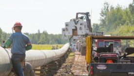 A construction worker in a red hardhat and blue shirt stands next to a white pipeline