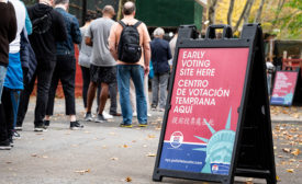 In the foreground is a red and blue sign with an image of the statue of liberty and text that reads, "early voting here." Behind it is a line of people on the sidewalk. 