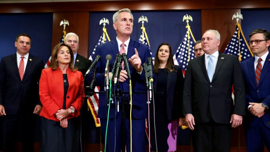 Senator McCarthy, wearing a blue suit and pink tie, stands at a press conference in front of 7 mics with a group of people standing in a loose horizontal line behind him. 