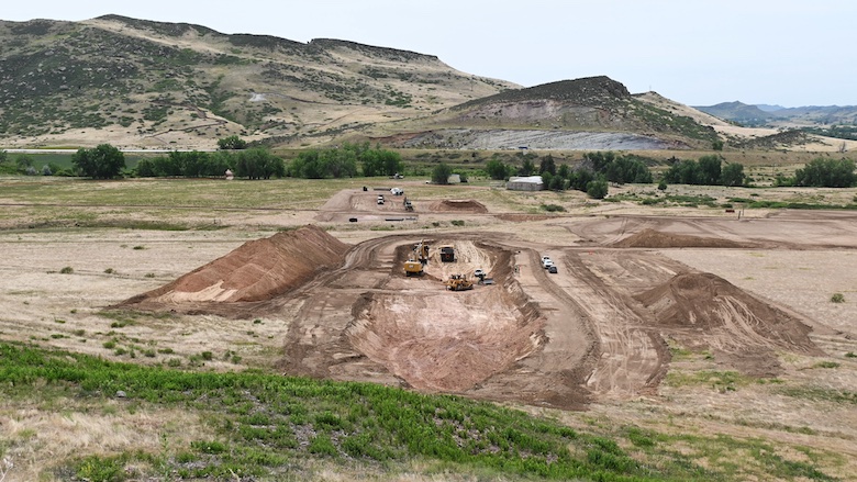 A construction site is shown in the foreground, with vehicles carving out a hole, with dirt mounds on either side. A row of bushes and mountains are in the background.  