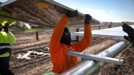 Two workers, one dressed in a bright orange jacket and another in neon yellow hold up a solar panel ready to be installed. You can see a reflection of the muddy dirt below in the underbelly of the panel. 