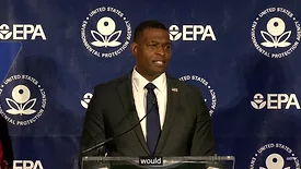 EPA administrator Michael Regan strands behind a translucent podium, wearing a black suit jacket, white collar shirt, navy blue tie and an American flag pin. The Navy blue background behind him is interspersed with white EPA logos. 