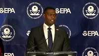 EPA administrator Michael Regan strands behind a translucent podium, wearing a black suit jacket, white collar shirt, navy blue tie and an American flag pin. The Navy blue background behind him is interspersed with white EPA logos. 