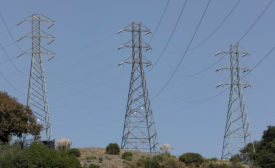 Three transmission towers stand in a row, spaced apart. A blue sky is in the background. 