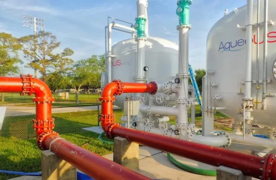 In this outdoor scene, orange red pipes shown in the foreground connect to two white water storage tanks. Trees and grass are seen in the background amidst a blue sky.