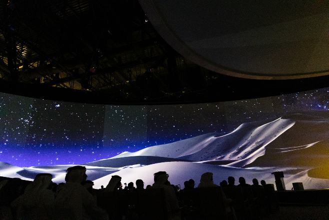 Silhouttes of attendees sit in chairs watching the image of an iceberg across a wide screen