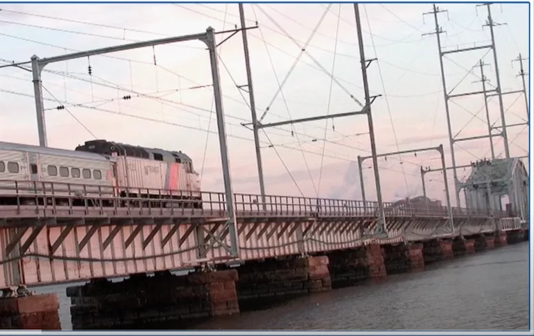 A New Jersey Transit train crosses over a bridge, with a pink and blue sky in the background.