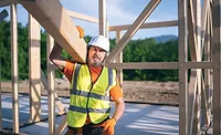 Construction worker wood frame A construction worker wearing an orange shirt and gloves, white hardhat and yellow vest holds up a wooden beam, with the frame of a structure in the background.