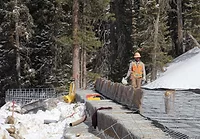a construction worker walks along a section of highway being rebuilt, with some snow on the ground and pine trees in the background