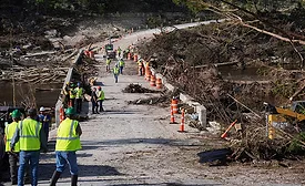 Workers wearing hardhats and yellow vests clean up in the aftermath of a flood, with debris seen on either side of a bridge