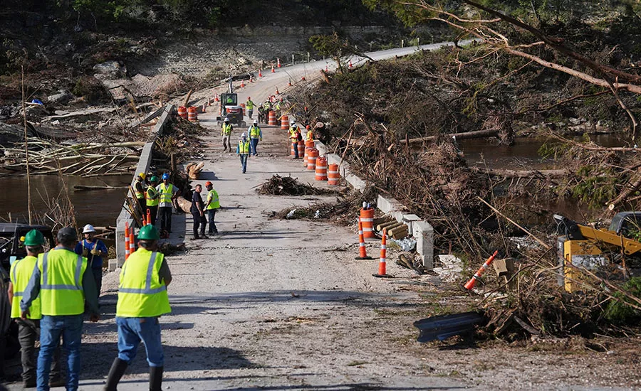 Workers wearing hardhats and yellow vests clean up in the aftermath of a flood, with debris seen on either side of a bridge