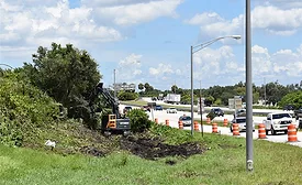 Excavator clears vegetation beside I-275 in Pinellas County, with live traffic and orange construction barrels visible in adjacent lanes.