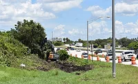 Excavator clears vegetation beside I-275 in Pinellas County, with live traffic and orange construction barrels visible in adjacent lanes.
