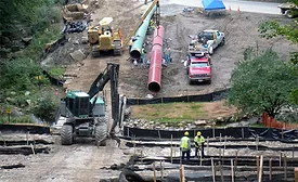 Workers and heavy equipment assemble large pipeline sections at a construction site with erosion-control barriers and wetland protections in place.