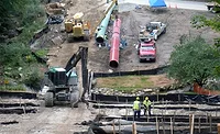 Workers and heavy equipment assemble large pipeline sections at a construction site with erosion-control barriers and wetland protections in place.