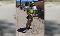 Utility worker wearing safety gear uses a listening device over a manhole cover on a gravel road to locate underground water infrastructure.