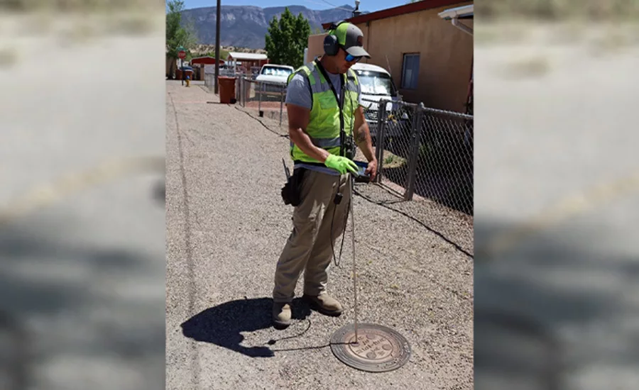 Utility worker wearing safety gear uses a listening device over a manhole cover on a gravel road to locate underground water infrastructure.