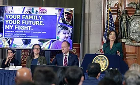 NY Governor Kathy Hochul stands in front of an audience as she presents the state budget at a press conference. A project screen on the stage reads