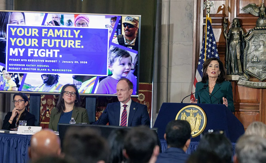 NY Governor Kathy Hochul stands in front of an audience as she presents the state budget at a press conference. A project screen on the stage reads