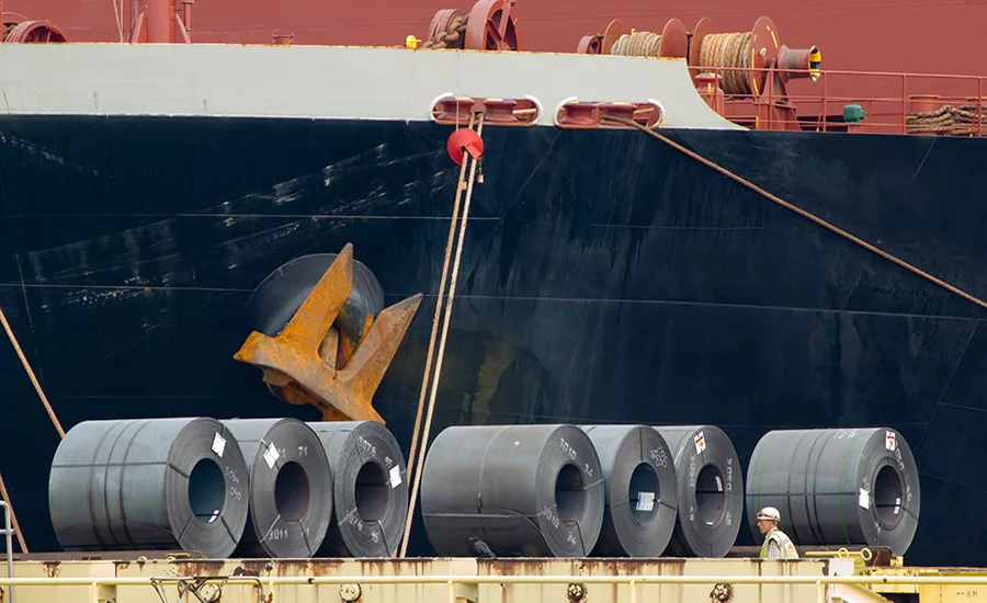 Steel coils unloaded at a port in front of a cargo vessel, illustrating tariff exposure on imported construction materials.