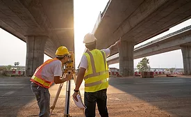 Construction workers using surveying equipment beneath highway overpass during infrastructure project work.