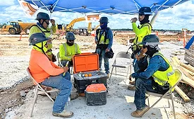 Construction workers rest under a shade canopy beside coolers and drinking water at an active jobsite during hot weather.