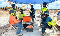 Construction workers rest under a shade canopy beside coolers and drinking water at an active jobsite during hot weather.