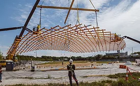 Photo shows a section of the timber gridshell being lifted into place by a crane, with one worker operating at height and another on the ground in the center of the photo