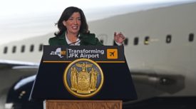 Gov. Hochul stands behind a podium wearing a green suit jacket and white dress shirt. A portion of a plane at JFK airport is visible behind her. 