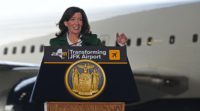 Gov. Hochul stands behind a podium wearing a green suit jacket and white dress shirt. A portion of a plane at JFK airport is visible behind her. 