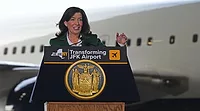 NYGovHochul.jpg Gov. Hochul stands behind a podium wearing a green suit jacket and white dress shirt. A portion of a plane at JFK airport is visible behind her.