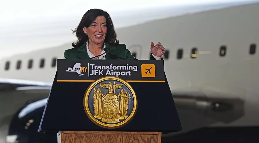 Gov. Hochul stands behind a podium wearing a green suit jacket and white dress shirt. A portion of a plane at JFK airport is visible behind her.
