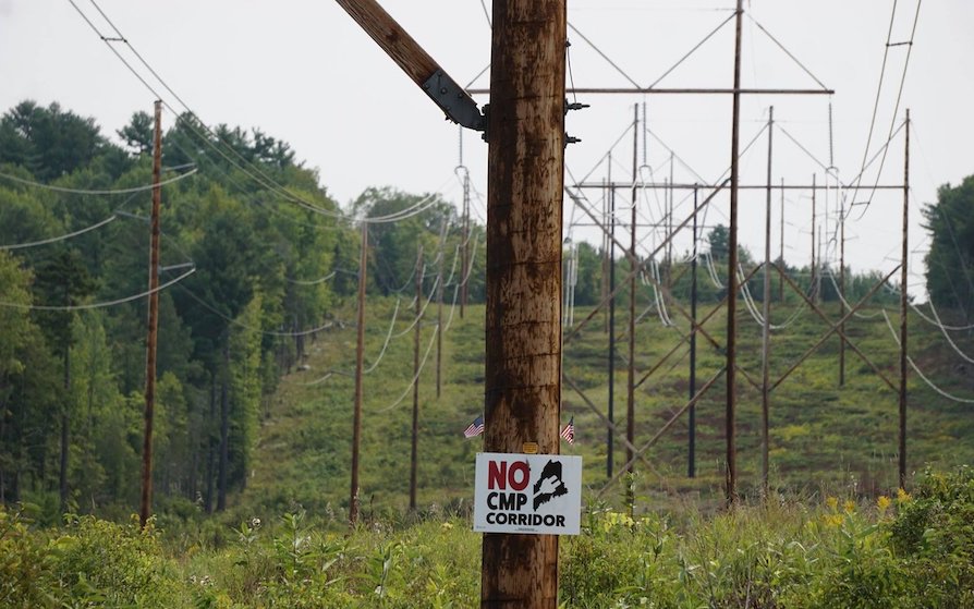 A sign posted on a pole centered among a green field, trees and power lines reads "No CMP Corridor"