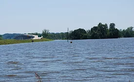 A body of flowing water is shown in the foreground. Behind it is a strip of green trees and a road with two cars