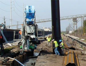 Workers use hydraulic-reaction press to drive steel sheet piles into difficult ground.